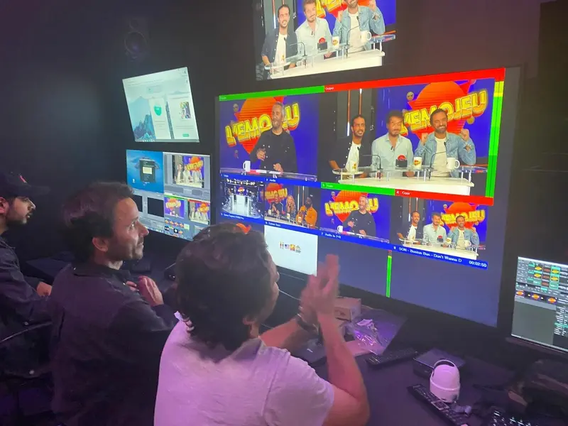 Des techniciens manipulent des mélangeurs vidéo et des moniteurs dans une régie, affichant plusieurs flux de caméras en direct d’une émission télévisée animée par trois présentateurs.