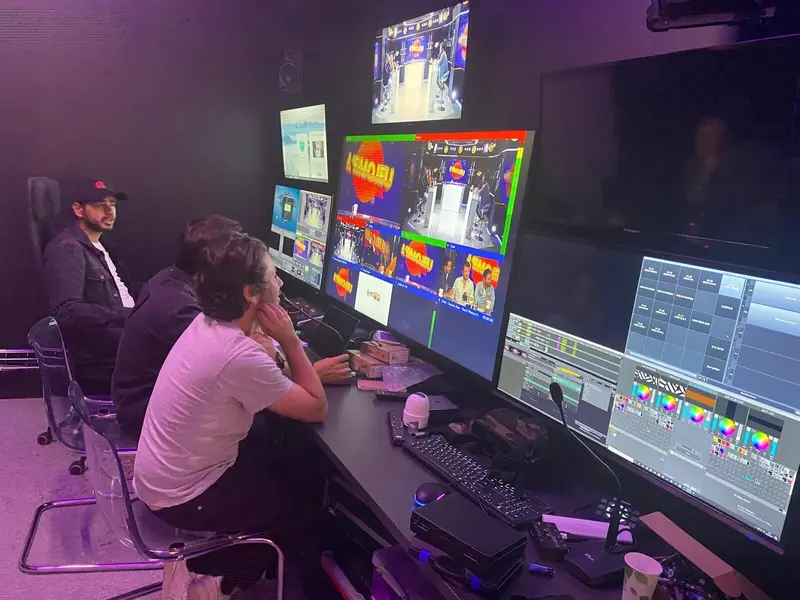 Trois personnes sont assises à un bureau de régie, surveillant plusieurs écrans affichant la production en direct d’un événement avec des graphismes dynamiques et des flux de caméras. La pièce, faiblement éclairée, est remplie d’équipements techniques et de moniteurs.