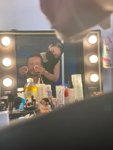 Un maquilleur portant un masque applique du maquillage à un homme assis devant un miroir de maquillage bien éclairé, avec divers produits et outils de maquillage disposés sur la table.