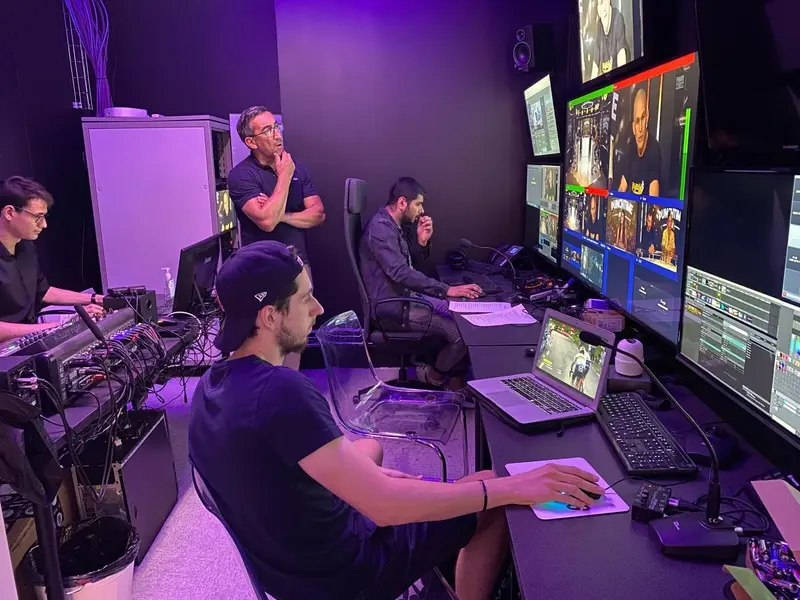 Quatre hommes travaillent dans une régie faiblement éclairée, entourés de plusieurs moniteurs, de flux vidéo et d’équipements techniques. L’un est assis à un bureau, utilisant une souris, tandis que les autres observent attentivement les écrans ou se tiennent debout à proximité.