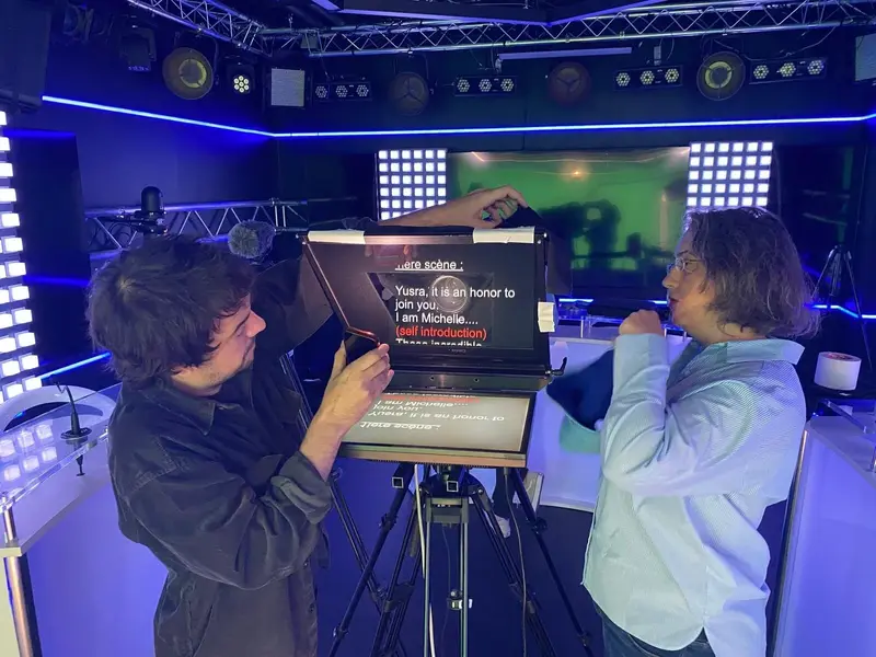 Deux personnes règlent un téléprompteur dans un studio très éclairé avec des lumières d'appoint bleues. Le téléprompteur affiche du texte et divers équipements et écrans sont visibles à l'arrière-plan.