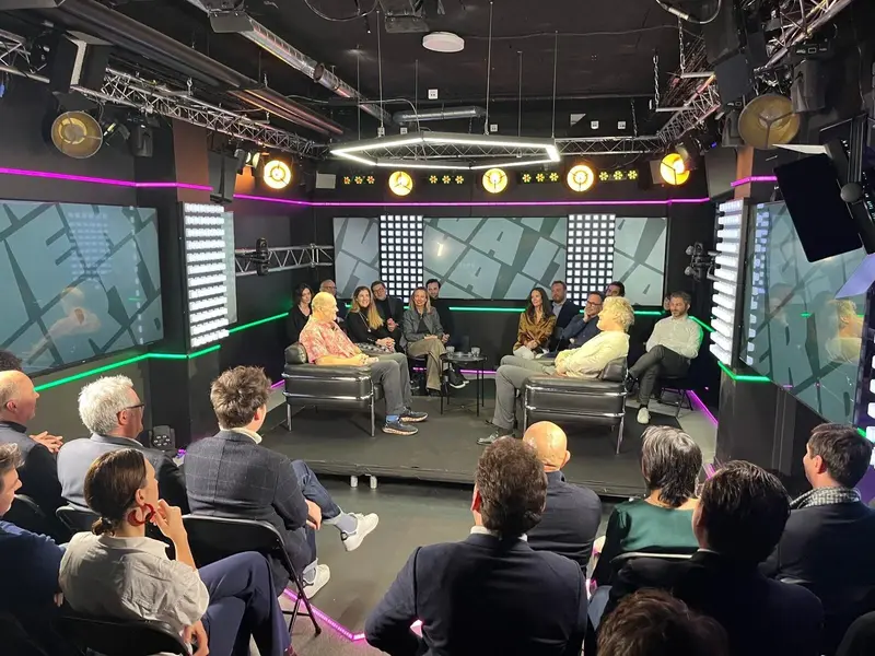 Un groupe de personnes est assis en demi-cercle sur une scène dans un studio moderne, entouré d’un public. Le décor, avec ses lumières vives et ses motifs géométriques, crée une atmosphère dynamique propice à la discussion.