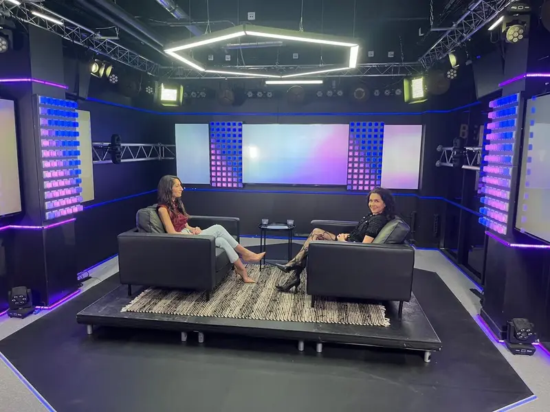 Deux femmes sont assises et discutent dans des fauteuils noirs dans un studio moderne et lumineux, avec un éclairage d’accent bleu et violet. Elles se font face, avec une petite table entre elles, sur un tapis noir et blanc.