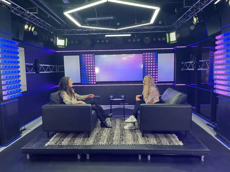 Deux femmes sont assises face à face dans des fauteuils noirs sur une petite scène, dans un studio moderne et lumineux avec des lumières colorées et un grand écran numérique en arrière-plan.