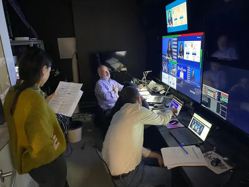 Trois personnes travaillent dans une salle de contrôle sombre équipée de plusieurs moniteurs et écrans d’ordinateur affichant des appels vidéo et des graphiques. Une personne est debout tenant des documents, tandis que deux autres sont assises à des bureaux avec ordinateurs portables et papiers.