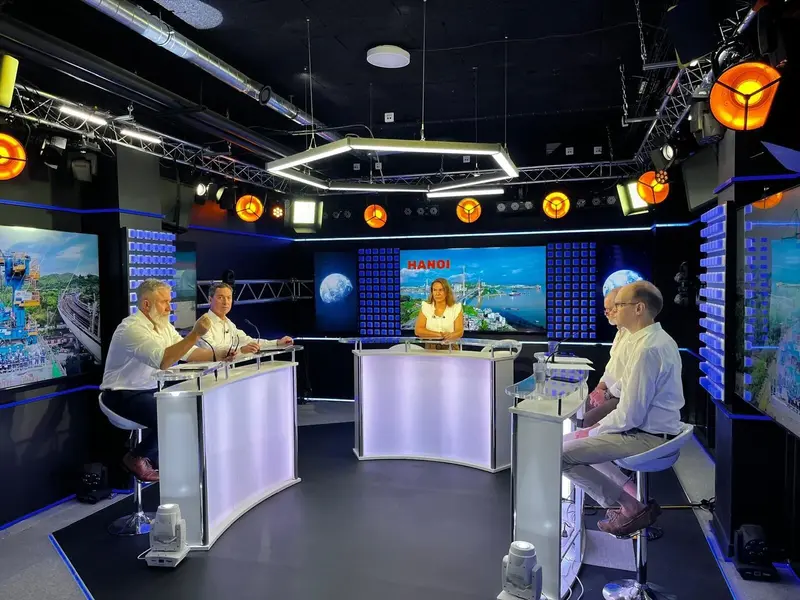 Cinq personnes sont assises à des bureaux modernes et éclairés dans un plateau de télévision, avec de grands écrans affichant des images de la ville et le mot HANOI en arrière-plan. Des projecteurs et du matériel de caméra sont visibles au-dessus.